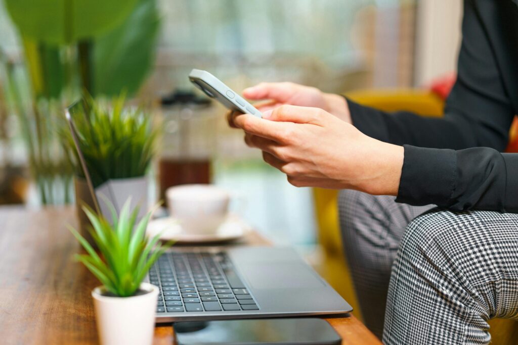 A person using a smartphone in a cozy home office setting with plants, a laptop, and coffee.